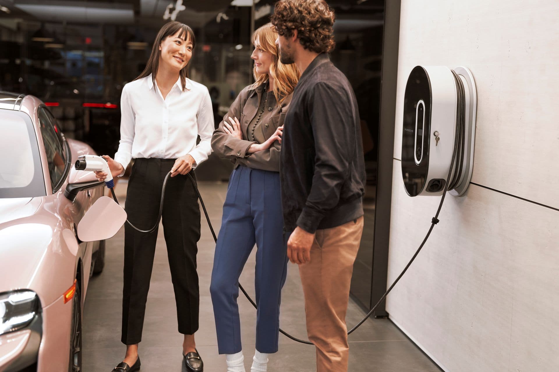 Three people are chatting beside a porsche taycan being charged at a charging station indoors.