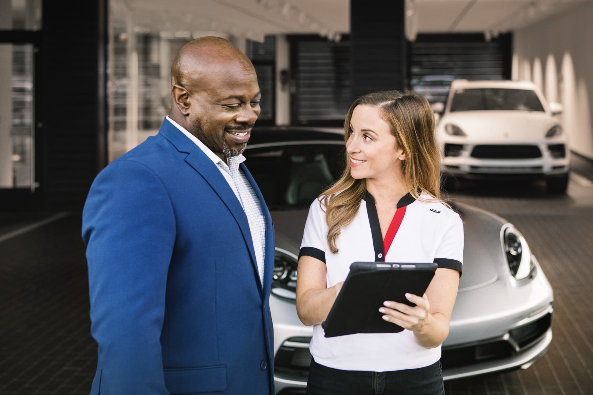 A man in a blue suit and a porsche service tech with a tablet smile while discussing inside a car showroom, with luxury cars in the background.