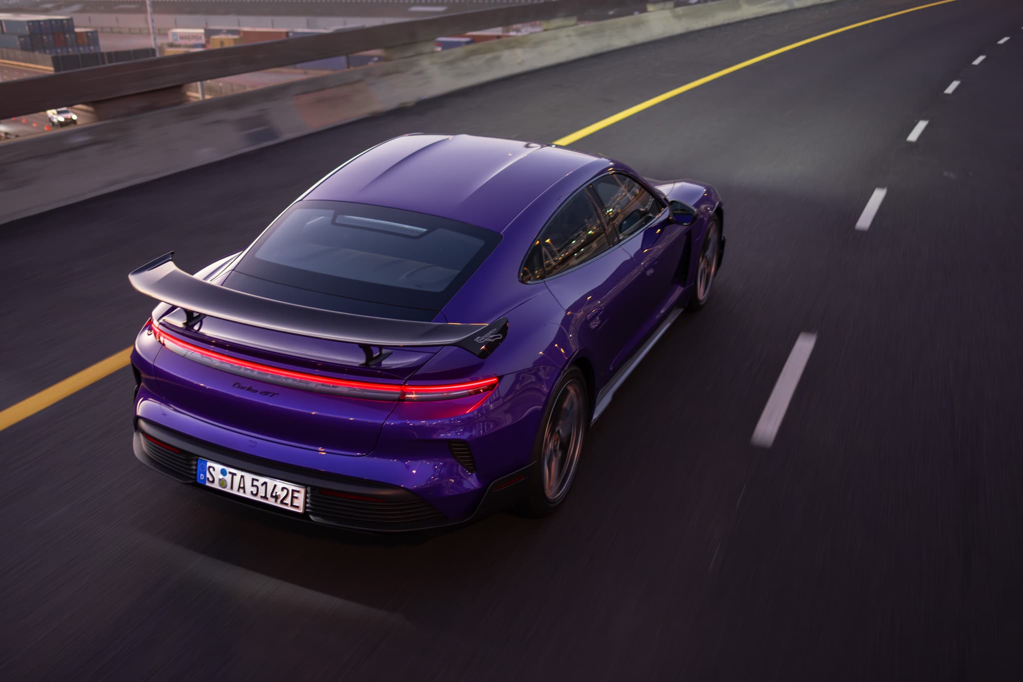 A purple porsche taycan with a spoiler drives on a highway, surrounded by a cityscape, during dusk.
