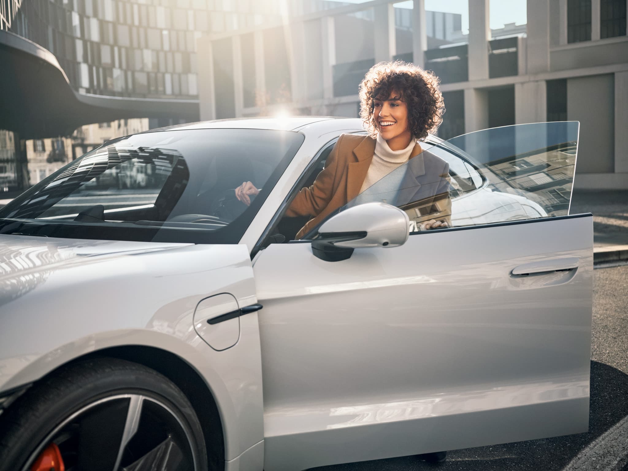 A person smiles while exiting a sleek white car on a sunny day, with modern buildings in the background.