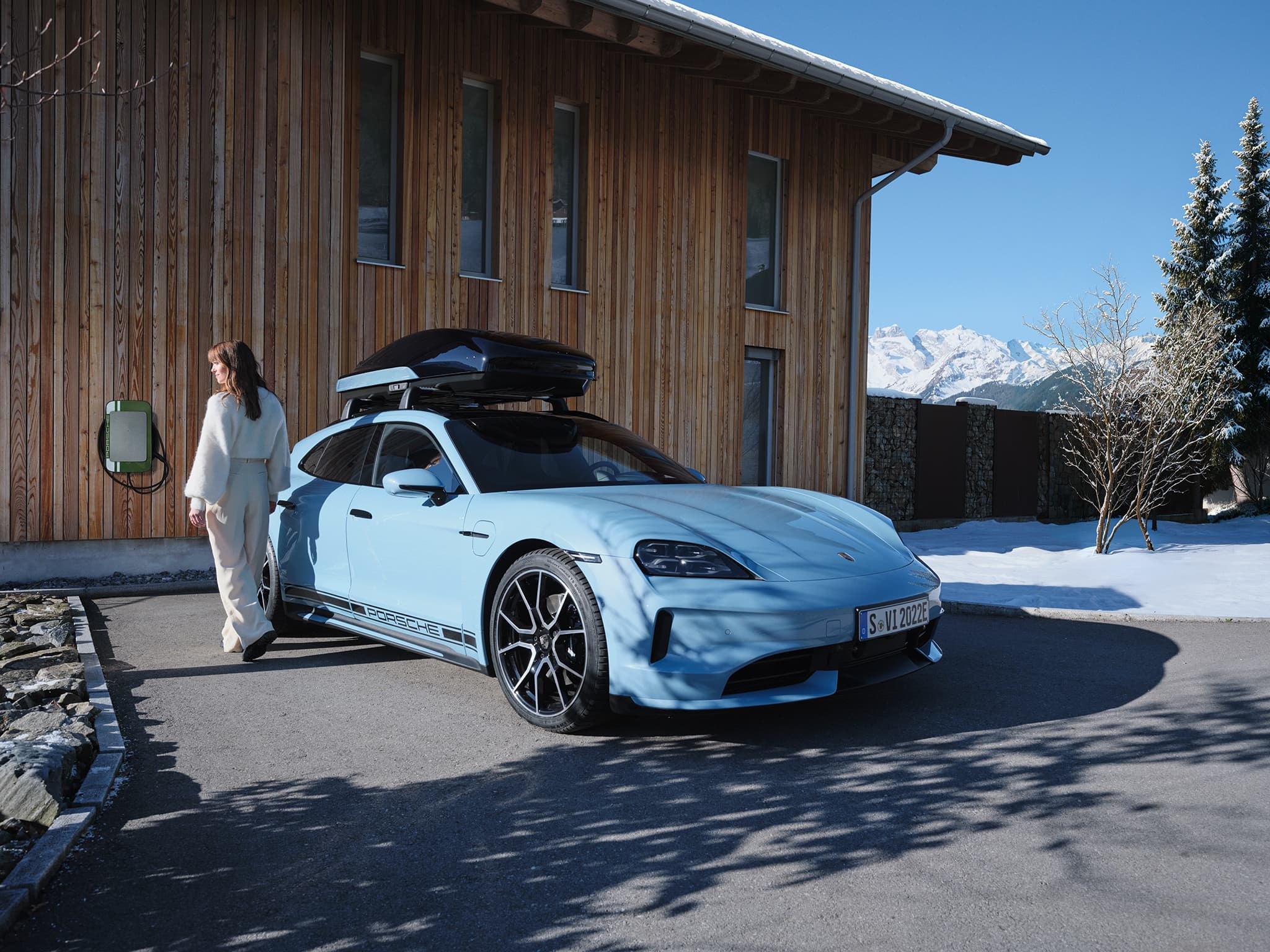 A woman walks beside a light blue porsche taycan with a roof box, parked near a wooden building in a snowy mountain setting.